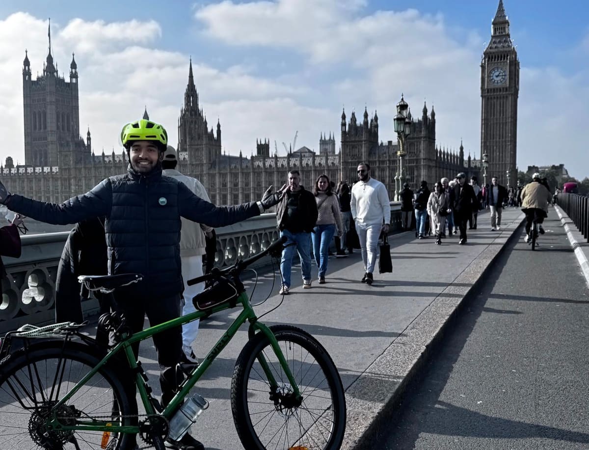 Sahil Jha with arms outstretched beside his bicycle on Westminster Bridge, with the Houses of Parliament and Big Ben behind him