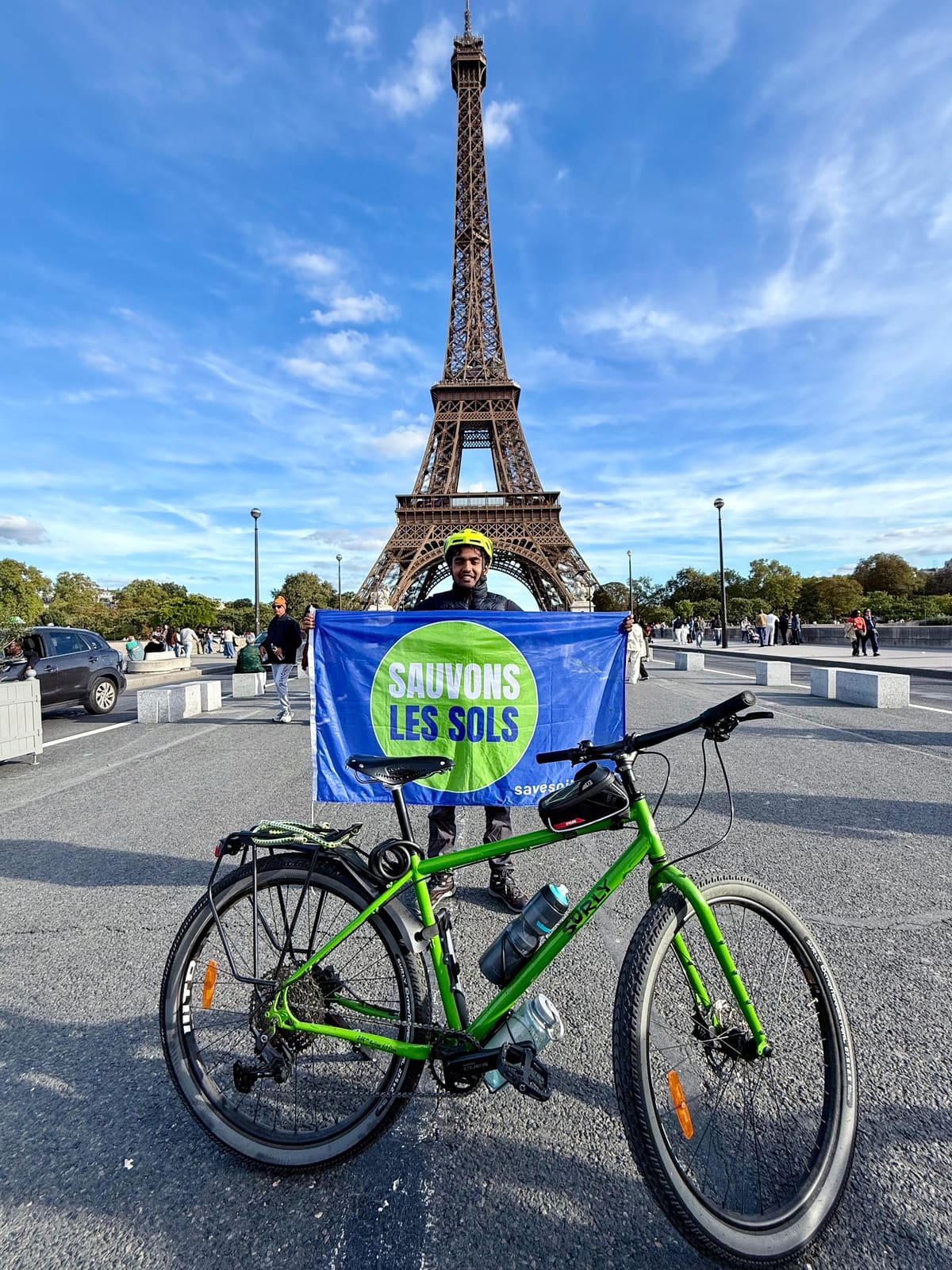Sahil Jha holding a "Sauvons les sols" Save Soil flag behind his green touring bicycle in front of the Eiffel Tower, Paris